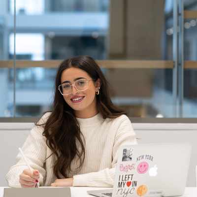 A woman with long hair and glasses sits at a desk smiling, using a stylus on a tablet. She is in front of a laptop with various stickers. Next to her, a mannequin displays a white T-shirt with colorful text. The setting is a modern office.