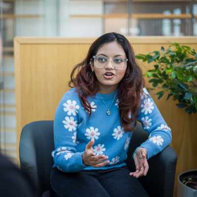 A young woman with glasses and long hair, wearing a blue sweater with white flower patterns, gestures while talking. She is seated in a modern indoor space, facing another person whose back is to the camera. A green plant is in the background.