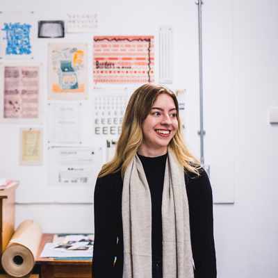 A person with long hair and a scarf stands smiling in an art studio. Behind them is a wall with various vintage posters and prints. A wooden chest of drawers and rolls of paper are visible in the foreground.