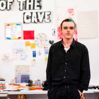 A person in a black shirt stands in a creative studio space. The wall behind them features a large OF THE CAVE sign and is covered with colorful artwork and posters. The overall atmosphere is artistic and vibrant.