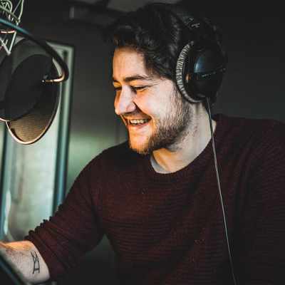 A man wearing headphones smiles while sitting in front of a microphone in a recording studio. He holds a pen and appears to be working or recording audio. The setting is dimly lit, with a lamp illuminating his workspace.