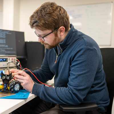 A person with glasses is seated at a desk in a lab, working on an electronic device with red wires. They are surrounded by equipment, including monitors and oscilloscopes. A whiteboard with writing is visible in the background.