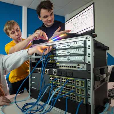 Three people work together on computer networking equipment. They are examining cables and a monitor displaying diagrams. The setting is a professional tech environment with a blue accent wall.