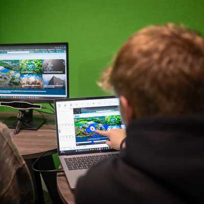 Two people are looking at computer screens in an office. One person points at a laptop displaying a design of a landscape, while a larger monitor shows similar images. The room has bright green walls.