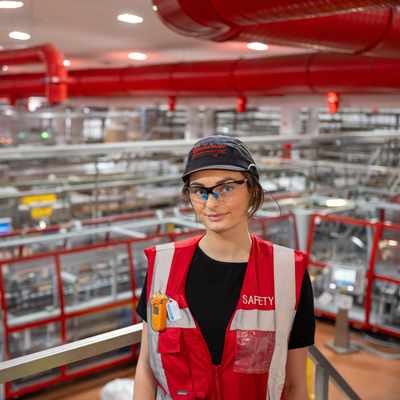 A person wearing safety gear and a cap stands in a large industrial facility with extensive machinery and red overhead pipes. The person is wearing a red vest labeled SAFETY and safety goggles, and looks toward the camera.