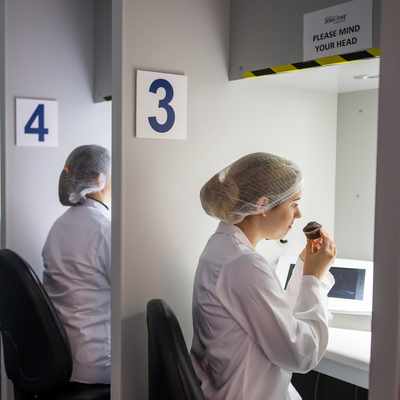 A row of people in white lab coats and hairnets sit at numbered stations. Each person is engaging in a sensory evaluation, with one person smelling or tasting from a small cup under a hooded light. The stations are numbered 3 to 8.