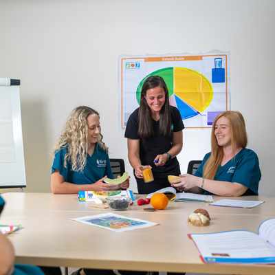 A group of women are seated at a table in a classroom setting, engaged in a discussion about nutrition. They have fruits on the table, and a pie chart on the wall is related to food groups. One woman is pouring a drink, while another peels a banana.