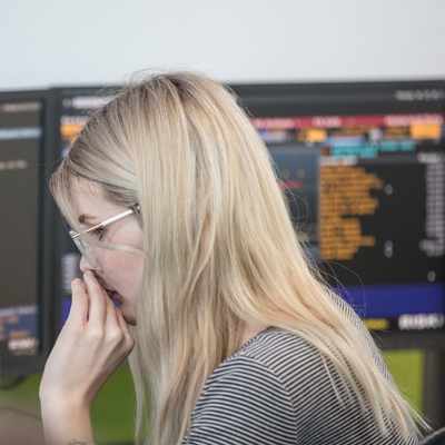 A woman with blonde hair and glasses is intently focused on monitors displaying complex data and graphs. She wears a striped shirt and appears to be in a busy office environment.