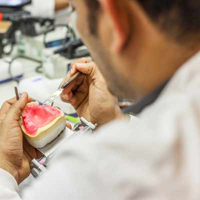 A dental technician works on a dental mold, meticulously shaping it with tools in a well-equipped lab. A small flame burner is visible on the table, suggesting a process that involves heat.