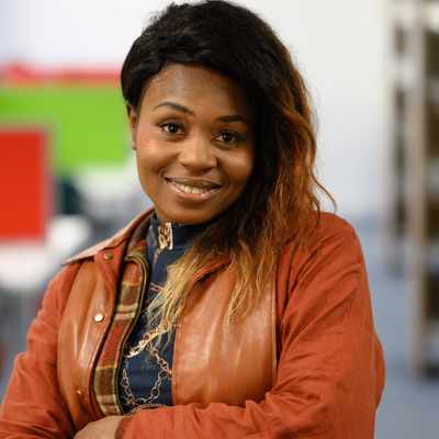 A woman with shoulder-length hair smiles warmly at the camera, standing in an office setting. She is wearing a brown jacket over a patterned top. The background features brightly colored partitioned workstations in red and green.