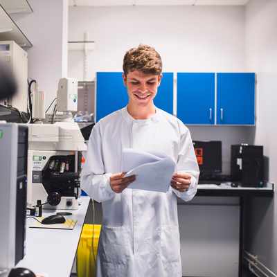 A smiling person in a lab coat holds papers in a laboratory with microscopes and computers. Shelves with blue cabinets are in the background. The environment suggests a research or scientific setting.