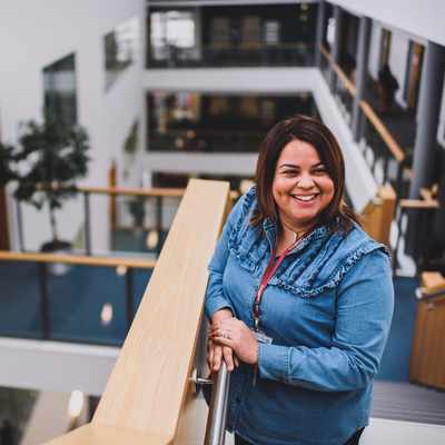 A woman smiles while leaning against a railing in a modern, multi-level office building. She is wearing a denim shirt and a lanyard. The background shows open hallways and glass barriers with indoor plants.