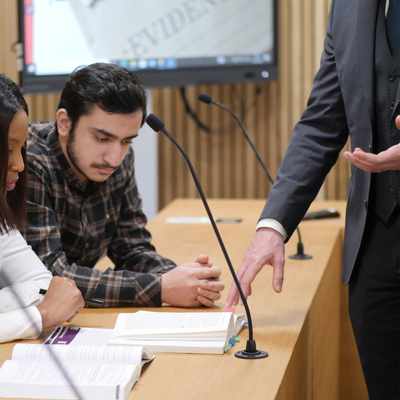 Four people are gathered around a table with open books and microphones, engaging in a discussion. A person in a suit stands nearby, gesturing with their hand. A screen in the background displays a blurred document with the word EVIDENCE.