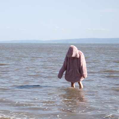 A person in a fluffy pink coat wades into the calm sea under a clear sky, with distant hills on the horizon.