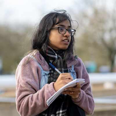 A person with long hair and glasses is outdoors, wearing a pink jacket and holding a notepad and pen. They seem to be taking notes or sketching. The background is blurred with trees and a structure visible.