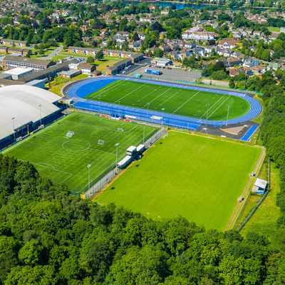 Aerial view of a sports complex with a blue athletics track surrounding a green football field. Adjacent is an indoor arena and another outdoor field, surrounded by lush trees and urban buildings in the background.