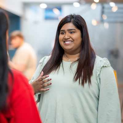 Two people are engaged in a conversation indoors. The person facing the camera is smiling and gesturing with their hand, wearing a light green top. The other person, with long hair, is turned away, wearing a red hoodie with white text.