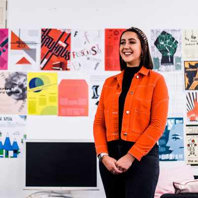 A person in an orange jacket stands smiling in front of a wall filled with colorful, abstract art posters. Theres a monitor and a couch in the foreground.