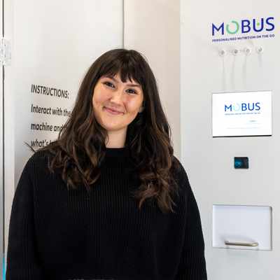 A person with long dark hair is smiling in front of a white vending machine labeled MOBUS. Text on the wall partially reads about understanding nutritional needs by 2035. The person is wearing a black sweater.