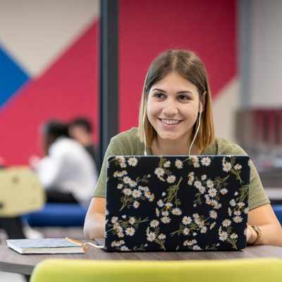 A woman smiling while using a laptop in a colorful, casual workspace. She is wearing earbuds and sitting at a round table. In the background, several people are gathered, and a foosball table is visible.
