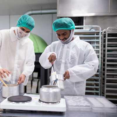 Two people in protective clothing and hairnets work in an industrial kitchen. One person holds a pot while the other pours ingredients into it. Stainless steel racks, equipment, and containers surround them.
