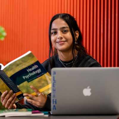 A person sits at a table with an open book titled Health Psychology and a laptop in front of them. They are smiling and positioned against a backdrop with red and orange vertical lines. Green plants are visible in the background.