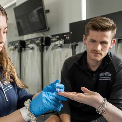 A woman in a blue hoodie and blue gloves is examining the hand of another person. A man in a black shirt observes the process. They are in a room with medical equipment visible in the background.