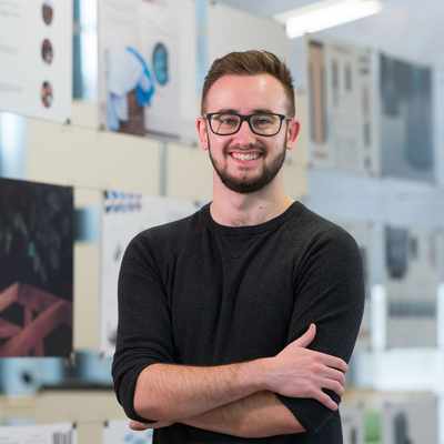 A man wearing glasses and a black sweater stands smiling with his arms crossed in front of display boards featuring various design projects and abstract images. The background is a well-lit room with natural light.