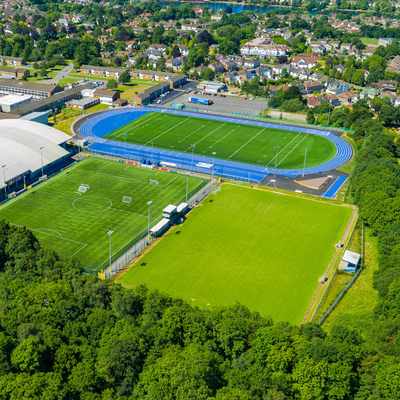 Aerial view of an athletic complex featuring a green soccer field, blue running track, and domed sports facility. Surrounding the complex are trees and residential buildings in the background.