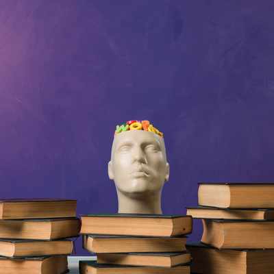 A mannequin head with colorful toy letters inside sits on a table surrounded by stacks of books against a purple background.
