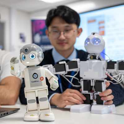 Two men interacting with robots in a tech setting. One is using a laptop, while two humanoid robots are on the table. The foreground robot is small and white with a delighted expression. The background has monitors and a blurred person.