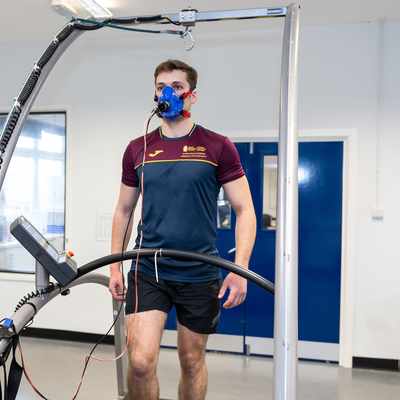 A man is walking on a treadmill wearing a blue mask for a fitness test. A woman sits nearby, operating a computer connected to the equipment. Both are in a lab setting wearing maroon and navy sports attire.