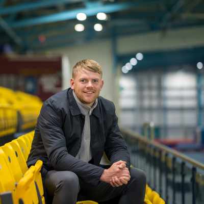 A man sits in an empty sports stadium with yellow seats, wearing a dark jacket and smiling. The background shows a brightly lit gymnasium interior with blue structural beams.