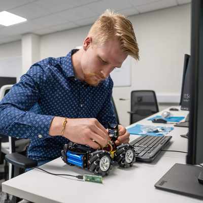 A person assembling a wheeled robot in an electronics lab, surrounded by tools and equipment.