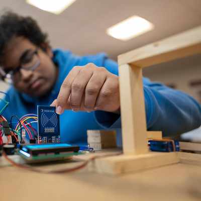A person in a blue hoodie works with electronic components and circuit boards, placing a small blue board among connecting wires on a table. The background is blurred and the lighting is bright.