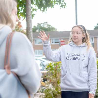 Young woman in grey hoodie and jeans gestures with hand while giving a campus tour to a group of visitors