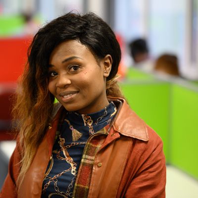 A person smiling and standing with arms crossed in a brightly colored office space. The background features red and green dividers and office furniture.