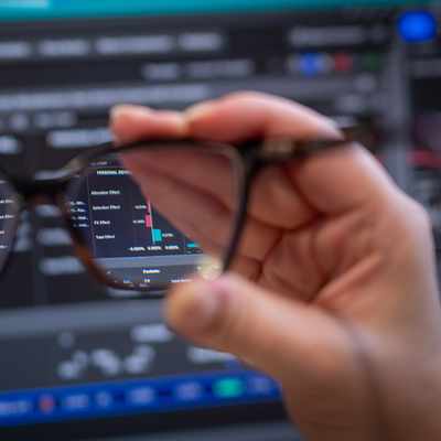 A woman with long blonde hair and glasses is concentrating in front of multiple computer screens displaying financial data. She is wearing a striped shirt and appears focused on her work.