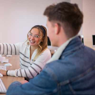 A person with short dark hair and glasses is smiling while sitting outdoors at a table using a laptop. They are wearing a green shirt, and the background is a blurry view of trees with sunlight filtering through.