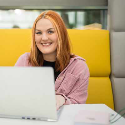 A woman with long brown hair and a striped shirt smiles while standing in an office. Theres a desk with a computer monitor and a plant in the background.