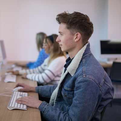 A man with glasses and a mustache is smiling while using a laptop outdoors. He is wearing a green shirt and is surrounded by a blurred, sunny background of trees and greenery.