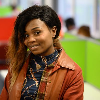 Two women are sitting and talking in an office setting. One woman with long hair and a patterned sweater smiles while the other, with hair tied back, faces her. Laptops are open on the table in front of them.