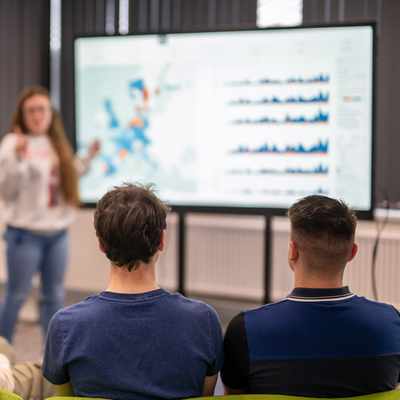Two people are focused on a computer screen, working together. The woman on the left is wearing a floral blouse, and the man on the right is in a black jacket. They are in a bright room with other computer equipment visible in the background.