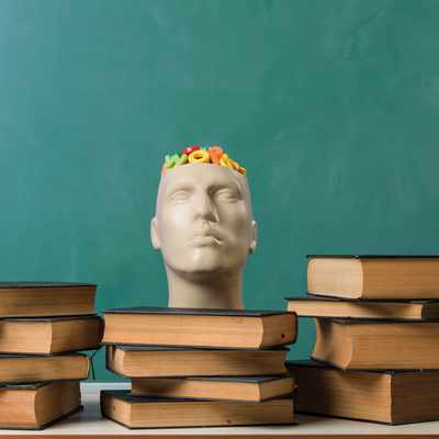 A person with curly hair is sitting at a table, smiling and writing in a notebook with a pen. They are wearing a gray sweater. The background features vertical wooden slats and a window.
