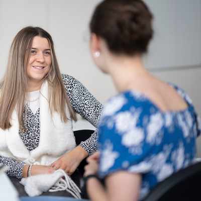 Two women sitting on a couch, one holding a clipboard and pen, smiling warmly at each other. The setting is a bright, cozy living room with a plant and artwork in the background.