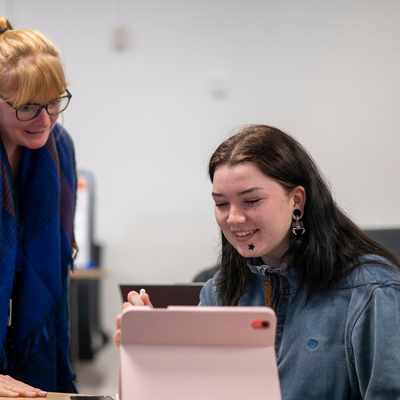 Two people are seated and talking at a table. One person in a green top and headscarf is smiling while using a laptop. The other, with curly hair, is seen from behind. They appear to be engaged in a positive conversation in an office setting.