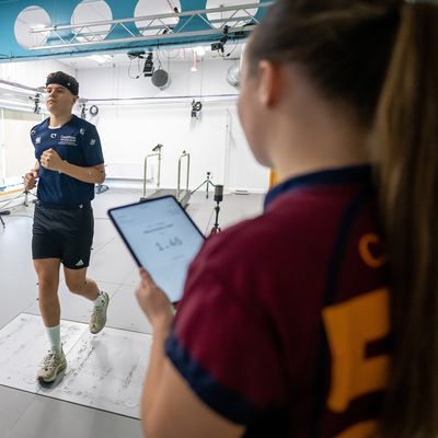 A person runs on a treadmill in a technology-equipped lab, surrounded by cameras and a computer screen. A bright green light is visible in the foreground. The setting suggests a motion analysis or biomechanics study.