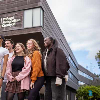 A group of four people walking and talking outdoors, holding coffee cups. Theyre dressed in casual winter clothing, with trees and modern buildings in the background under a partly cloudy sky.