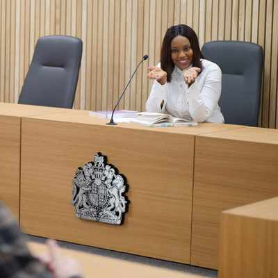 A man in a light gray suit and white shirt is sitting at a table, smiling and gesturing with his hands. He faces a person with long hair, seated across from him. A glass of water and papers are on the wooden table.