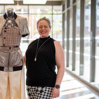 A person is focused on working with fabric in a fashion design studio. They are surrounded by dress forms and sewing materials, with others working in the background.